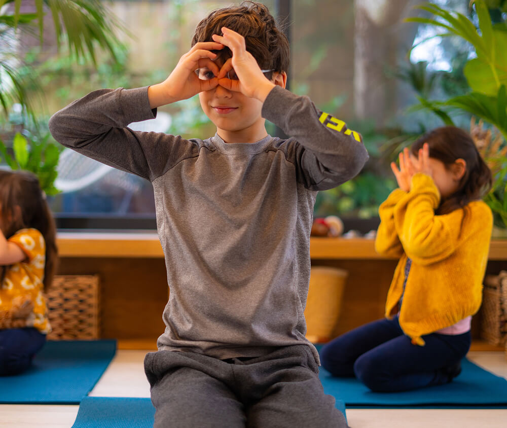 A child practicing Yoga on a blue mat with other kids in the background doing yoga